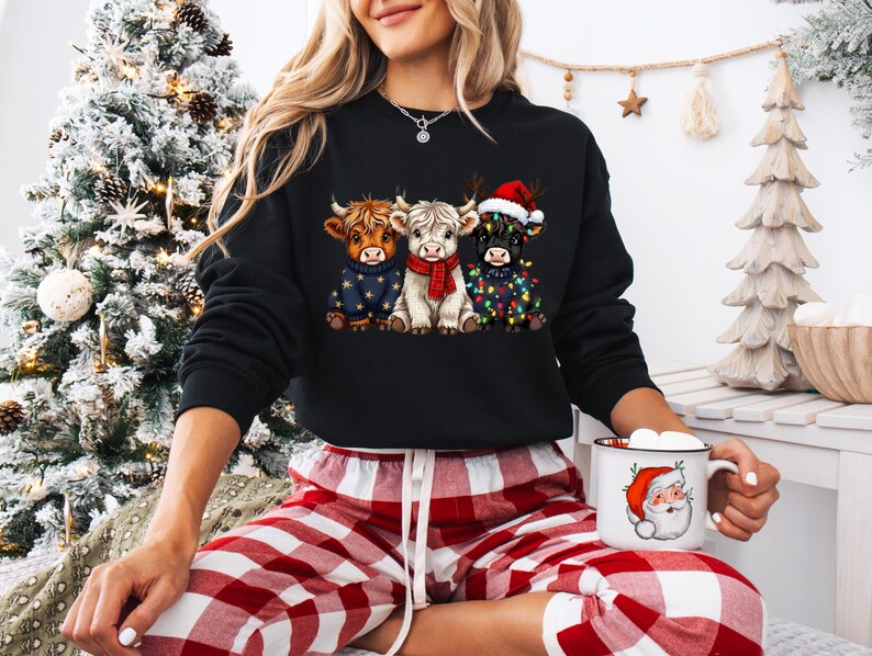 May include: Black sweatshirt featuring three Highland cows in festive attire. One cow wears a Santa hat, another is adorned with Christmas lights, and the third is in a blue sweater. The model is wearing red and white plaid pajama pants and holding a Santa mug.