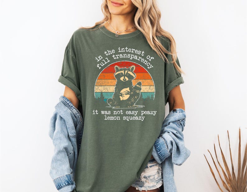 May include: Olive green t-shirt featuring a raccoon illustration within a retro sunset design. The text reads "in the interest of full transparency" and "it was not easy peazy lemon squeezy".