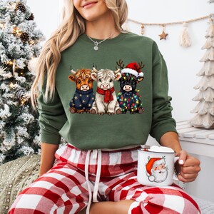 May include: An olive green sweatshirt featuring three Highland cows in festive attire. One cow wears a star-patterned sweater, another a red scarf, and the third a Santa hat and Christmas lights. The person is holding a Santa mug.