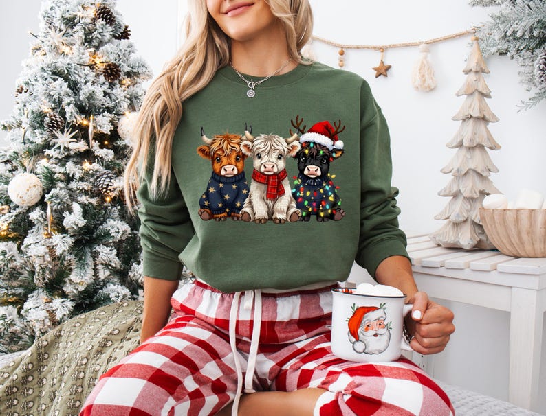 May include: An olive green sweatshirt featuring three Highland cows in festive attire. One cow wears a star-patterned sweater, another a red scarf, and the third a Santa hat and Christmas lights. The person is holding a Santa mug.