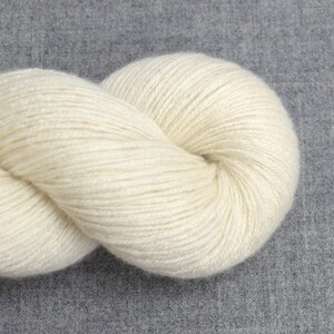 May include: A skein of off-white yarn, tightly wound and twisted, rests against a gray fabric background. The yarn appears soft and textured, ideal for knitting or crocheting projects. The neutral color makes it versatile for various craft projects.