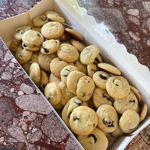 Puede incluir: Una caja blanca rebosante de galletas con chispas de chocolate recién horneadas. Las galletas son de color dorado con chispas de chocolate visibles. La caja está abierta, revelando una generosa porción de estas delicias.