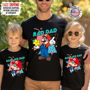 May include: Black t-shirts with a graphic of a cartoon character wearing sunglasses and a red hat. The shirts read "RAD DAD" and "RAD LIKE DAD" in a retro font. The shirts are worn by a father and two children.