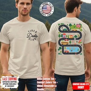 May include: Ivory-colored t-shirt with the text "Tired Dads Club" in black script. The back of the shirt features a colorful road design with cars, houses, trees, and a pond. Made in the USA.