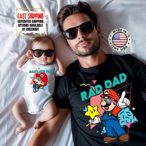 May include: A father and baby wearing sunglasses and matching graphic shirts. The baby wears a white onesie with "RAD LIKE DAD" and a cartoon character. The father's black shirt says "RAD DAD" with a similar cartoon design. Made in the USA.