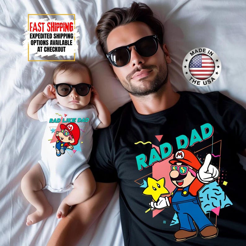 May include: A father and baby wearing sunglasses and matching graphic shirts. The baby wears a white onesie with "RAD LIKE DAD" and a cartoon character. The father's black shirt says "RAD DAD" with a similar cartoon design. Made in the USA.
