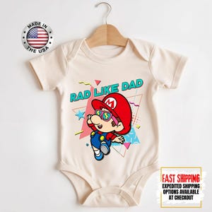 May include: Cream-colored baby bodysuit featuring a cartoon character in a red hat and sunglasses, with the text "RAD LIKE DAD." The design includes geometric shapes in pink, blue, and yellow. A "Made in the USA" emblem is visible.