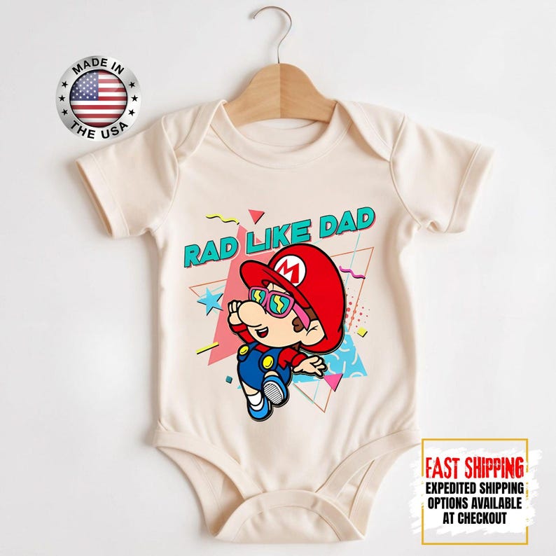 May include: Cream-colored baby bodysuit featuring a cartoon character in a red hat and sunglasses, with the text "RAD LIKE DAD." The design includes geometric shapes in pink, blue, and yellow. A "Made in the USA" emblem is visible.