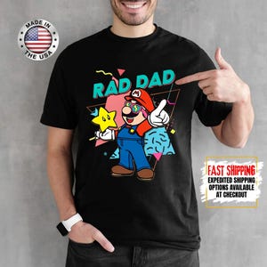 May include: Black t-shirt featuring a cartoon Mario character with the text "RAD DAD". The character is wearing sunglasses and holding a star. The shirt has a retro 80s style design.