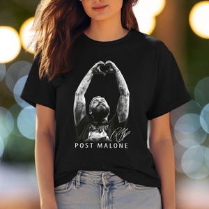 May include: A black short-sleeved t-shirt featuring a monochromatic graphic of Post Malone with arms raised to form a heart shape above the head. The design includes a signature and the text POST MALONE printed in white capital letters below the image.