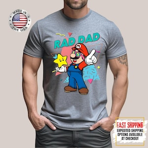 May include: Gray t-shirt featuring a cartoon illustration of a character with the text "RAD DAD". The character is wearing a red hat, sunglasses, and blue overalls, holding a star. The shirt has a "Made in the USA" logo.