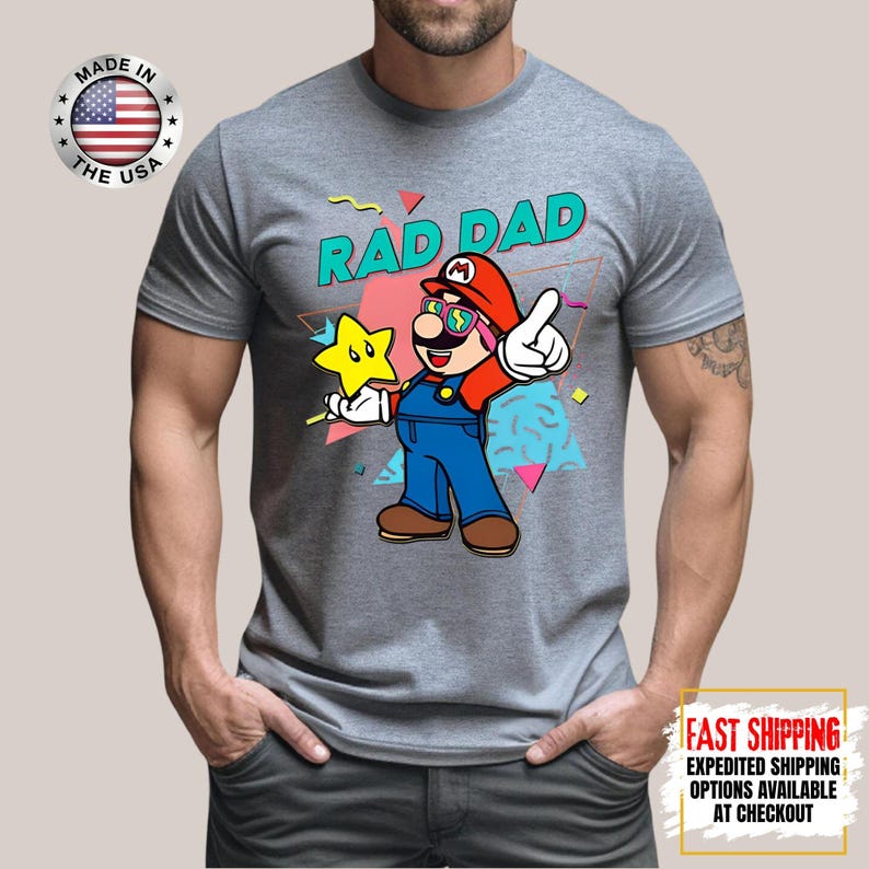 May include: Gray t-shirt featuring a cartoon illustration of a character with the text "RAD DAD". The character is wearing a red hat, sunglasses, and blue overalls, holding a star. The shirt has a "Made in the USA" logo.