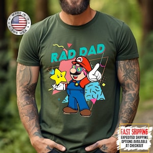 May include: Green t-shirt with a graphic of a cartoon character wearing sunglasses and a red hat, holding a yellow star. The text "RAD DAD" is above the character. The shirt also has a "Made in the USA" logo.