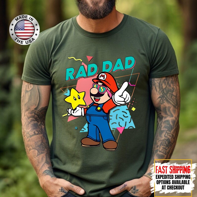 May include: Green t-shirt with a graphic of a cartoon character wearing sunglasses and a red hat, holding a yellow star. The text "RAD DAD" is above the character. The shirt also has a "Made in the USA" logo.