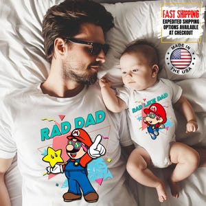 May include: A white t-shirt with "RAD DAD" and a cartoon character wearing sunglasses, a red hat, and blue overalls. A baby in a white onesie with "RAD LIKE DAD" and a matching cartoon character. The image includes "MADE IN THE USA" and "FAST SHIPPING" text.