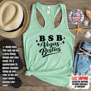May include: A light green tank top with a racerback design. The text "BSB Vegas Besties" is printed in black script. The tank top is displayed on a wooden surface with a straw hat, flip-flops, and sunglasses.