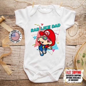May include: White baby bodysuit with a colorful graphic of a cartoon character wearing a red hat and sunglasses. The text "RAD LIKE DAD" is above the character. A "Made in the USA" sticker is visible.