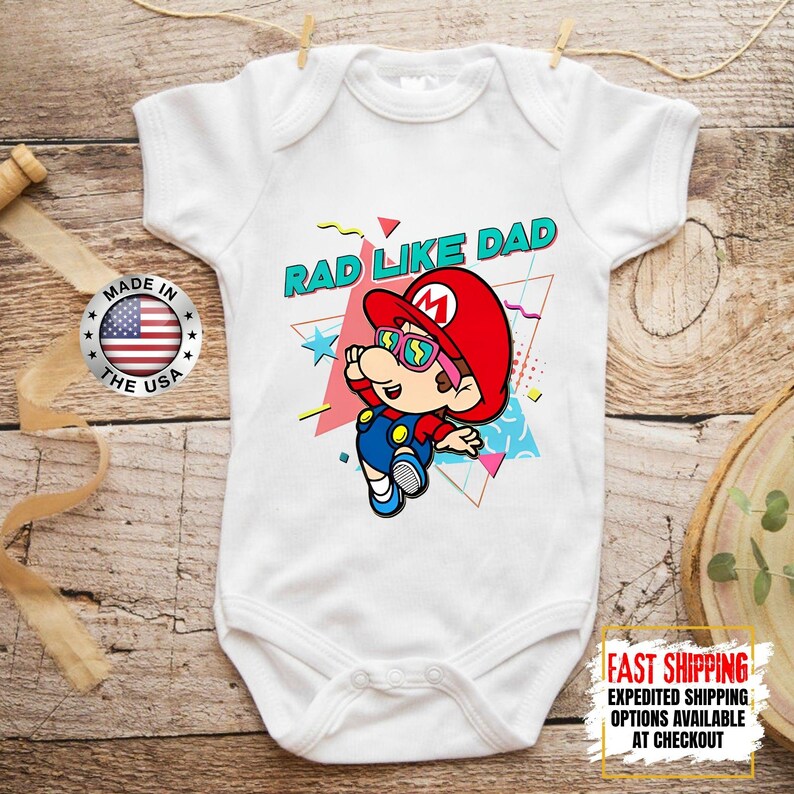 May include: White baby bodysuit with a colorful graphic of a cartoon character wearing a red hat and sunglasses. The text "RAD LIKE DAD" is above the character. A "Made in the USA" sticker is visible.