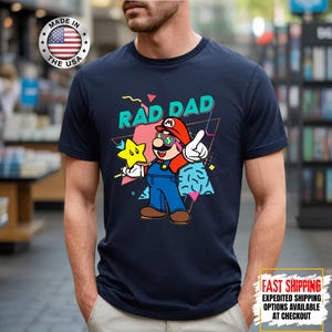 May include: Navy blue t-shirt featuring a cartoon illustration of a character with the text "RAD DAD". The character is wearing a red hat, sunglasses, and blue overalls, holding a star. The shirt has a "Made in the USA" logo.