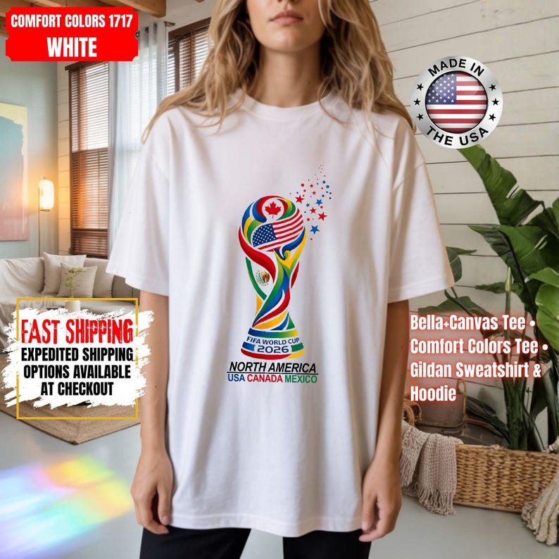 May include: A white short-sleeve t-shirt featuring a colorful graphic of the FIFA World Cup 2026 trophy. The trophy design incorporates the flags of the USA, Canada, and Mexico, with small red and blue stars above it and the text North America USA Canada Mexico.