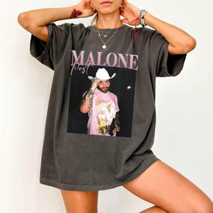 May include: A charcoal gray short-sleeve t-shirt featuring a graphic print of a person wearing a white cowboy hat and a pink shirt. The text MALONE is printed in large pink letters above the image, with the word Post written in a cursive script.