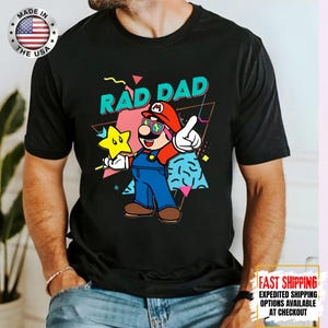 May include: Black t-shirt with a colorful graphic of a cartoon character wearing sunglasses and overalls, holding a star, with the words "RAD DAD". The shirt has a "Made in the USA" logo.