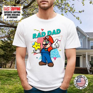 May include: White t-shirt featuring a cartoon illustration of a character in a red hat and overalls, holding a star. The text "RAD DAD" is above the character. The shirt has a colorful, retro design.
