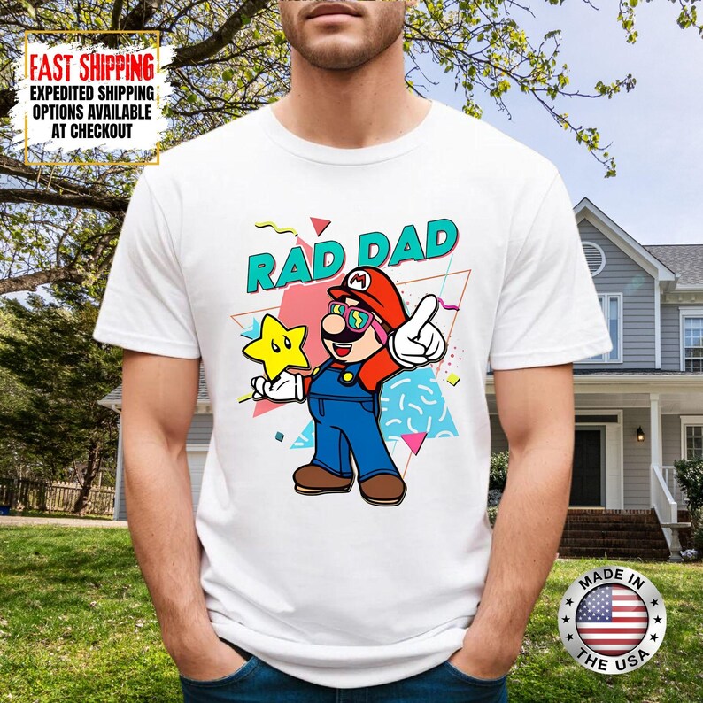 May include: White t-shirt featuring a cartoon illustration of a character in a red hat and overalls, holding a star. The text "RAD DAD" is above the character. The shirt has a colorful, retro design.