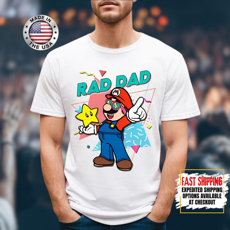 May include: White t-shirt featuring a cartoon illustration of a character with a red hat, sunglasses, and blue overalls, holding a yellow star. The text "RAD DAD" is above the character. A "Made in the USA" logo is visible.