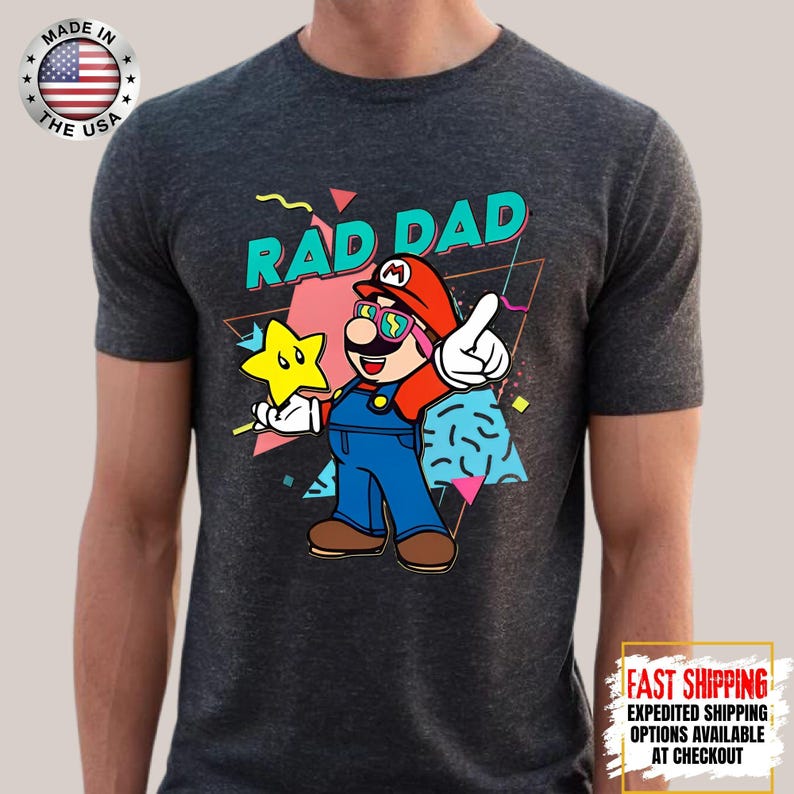 May include: Dark gray t-shirt featuring a cartoon illustration of a character with the text "RAD DAD". The character is wearing a red hat, blue overalls, and sunglasses, holding a yellow star. The shirt has a retro design.