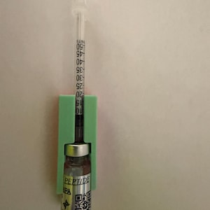 May include: A clear syringe with a scale from 10 to 50 units is inserted into a green holder, which also holds a small glass vial. The vial has a silver label with the word "PEPTIDE" and a QR code.