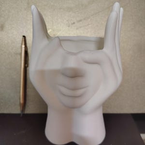 May include: A white ceramic planter shaped like a pair of hands cupping a space. The planter has a smooth finish and a detailed design. A gold-colored mechanical pencil is next to the planter.