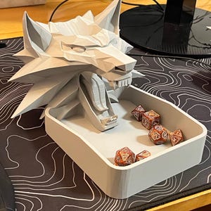 May include: A light gray wolf-shaped dice tray with a rectangular base. The wolf's head is detailed with sharp angles and pointed ears. Several multi-sided dice with red and gold patterns are inside the tray. The tray sits on a black and white patterned surface.