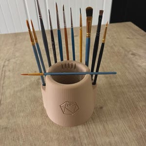 May include: A beige 3D-printed brush holder with a geometric design, holding various paint brushes with blue, black, and gold handles. The brushes are of different sizes and shapes, suitable for painting and art projects.