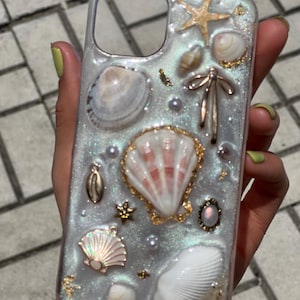 May include: Clear phone case decorated with seashells, pearls, and gold accents. The case features a variety of shells, including a starfish, scallop shells, and small conch shells, all set against a shimmering, pearlescent background.