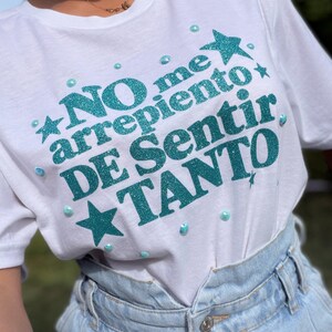 May include: A white t-shirt featuring the teal glitter text NO me arrepiento DE SENTIR TANTO, accented with teal glitter stars and small iridescent circular embellishments scattered across the front of the garment.