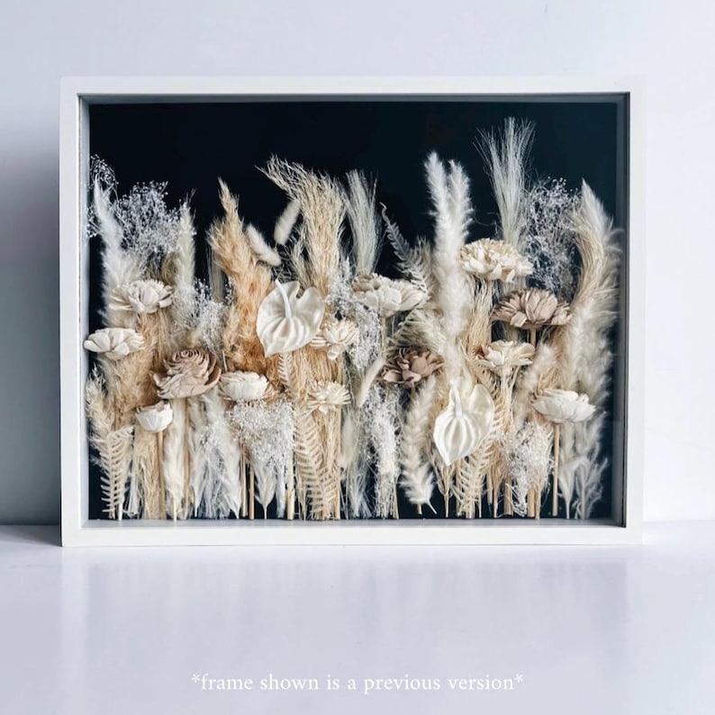 May include: A framed floral arrangement featuring dried flowers in shades of cream, beige, and brown against a black background. The arrangement includes various textures and shapes, with a white frame. The text at the bottom reads "frame shown is a previous version".