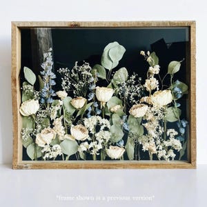 May include: A rectangular wooden framed floral arrangement featuring dried white roses, blue delphiniums, and green eucalyptus leaves against a black background. The frame is a light brown color. The text at the bottom reads "frame shown is a previous version".