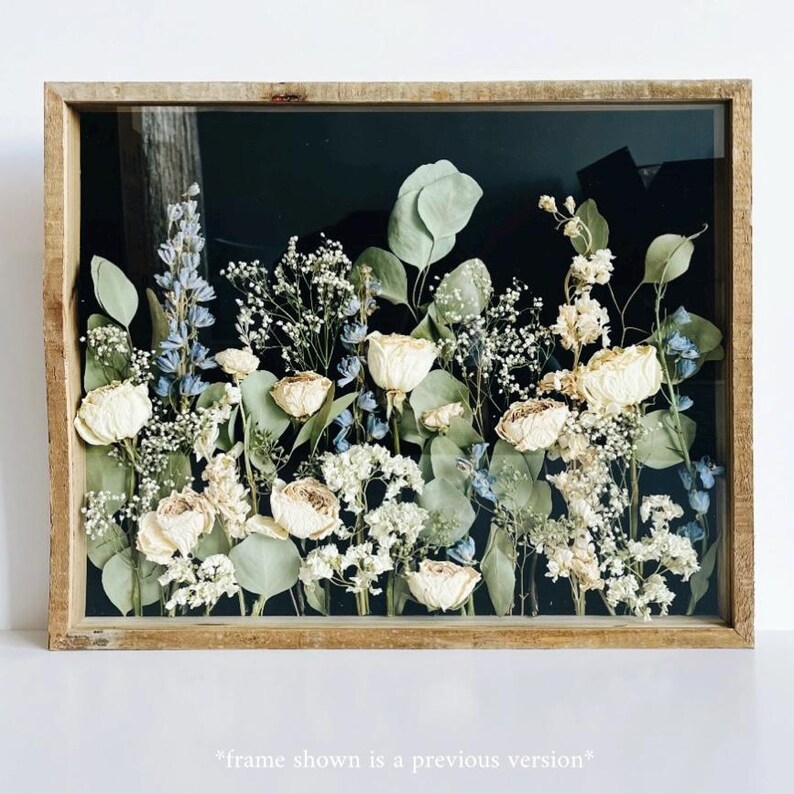 May include: A rectangular wooden framed floral arrangement featuring dried white roses, blue delphiniums, and green eucalyptus leaves against a black background. The frame is a light brown color. The text at the bottom reads "frame shown is a previous version".