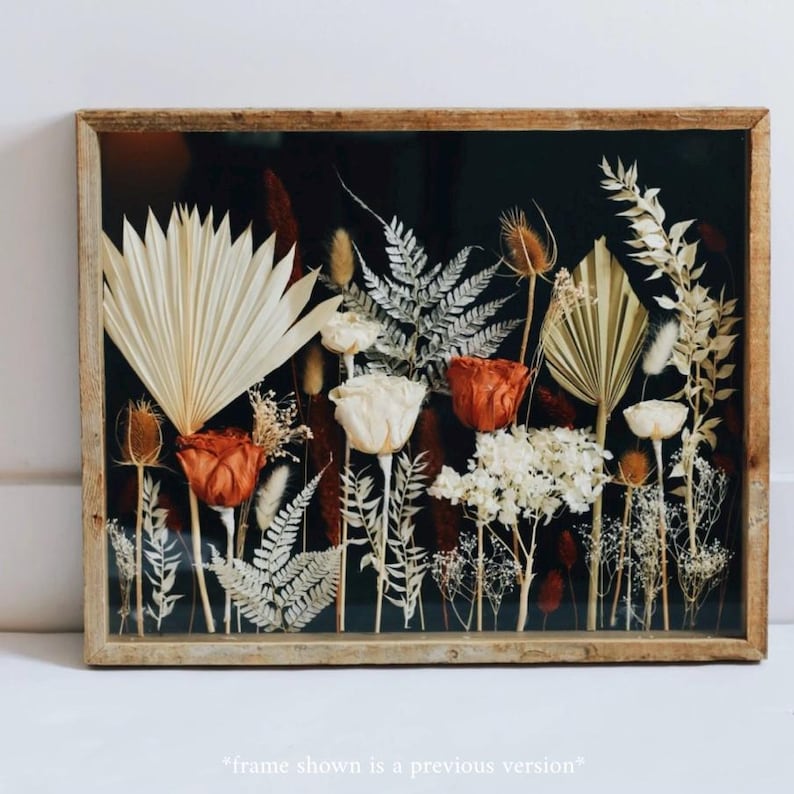 May include: A framed botanical art piece featuring dried flowers and foliage. The arrangement includes orange roses, white hydrangeas, and various dried grasses and ferns, set against a dark background. The frame is made of light-colored wood. The text at the bottom reads "frame shown is a previous version".