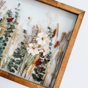 May include: A framed floral arrangement featuring dried flowers and greenery. The arrangement includes white cosmos flowers, eucalyptus, and various dried grasses and seed pods. The frame is made of stained wood, and the background is a soft white.