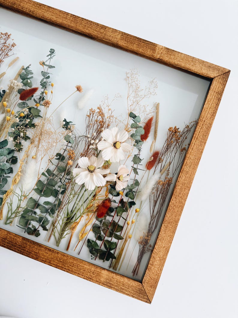 May include: A framed floral arrangement featuring dried flowers and greenery. The arrangement includes white cosmos flowers, eucalyptus, and various dried grasses and seed pods. The frame is made of stained wood, and the background is a soft white.