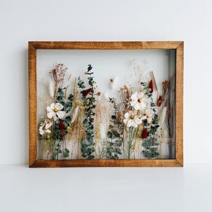 May include: A rectangular wooden framed piece of art featuring a display of dried flowers and foliage. The arrangement includes white flowers, green eucalyptus, and various dried grasses and seed pods. The frame is a warm brown color.