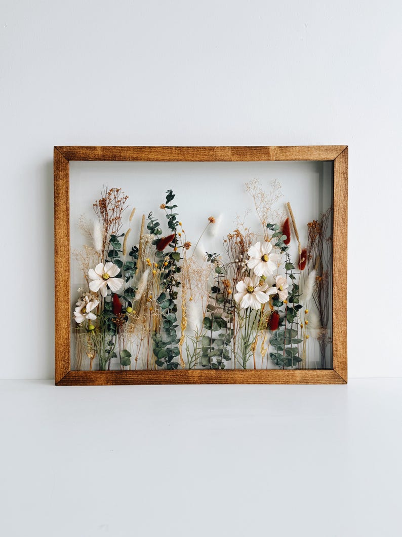 May include: A rectangular wooden framed piece of art featuring a display of dried flowers and foliage. The arrangement includes white flowers, green eucalyptus, and various dried grasses and seed pods. The frame is a warm brown color.