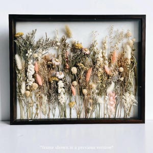 May include: A framed botanical art piece featuring a variety of dried flowers and grasses in shades of cream, pink, yellow, and brown. The arrangement is set against a white background within a dark brown wooden frame. The text at the bottom says "frame shown is a previous version".