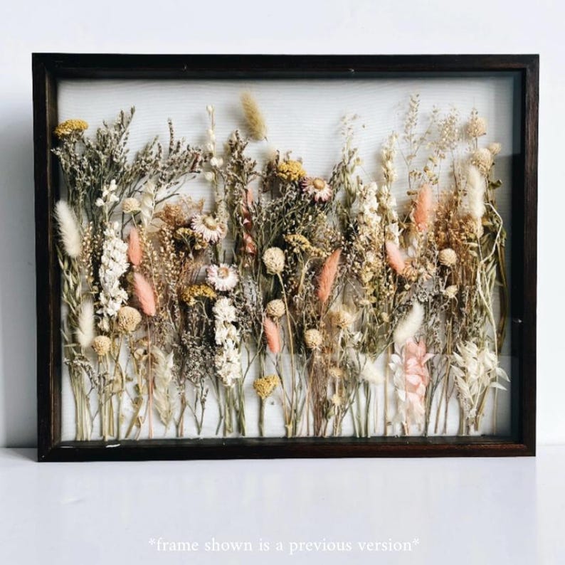 May include: A framed botanical art piece featuring a variety of dried flowers and grasses in shades of cream, pink, yellow, and brown. The arrangement is set against a white background within a dark brown wooden frame. The text at the bottom says "frame shown is a previous version".