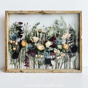 May include: A framed botanical art piece featuring dried flowers and foliage. The arrangement includes yellow roses, blue thistles, and burgundy leaves, set against a clear background. The frame is made of light-colored wood, creating a rustic aesthetic.