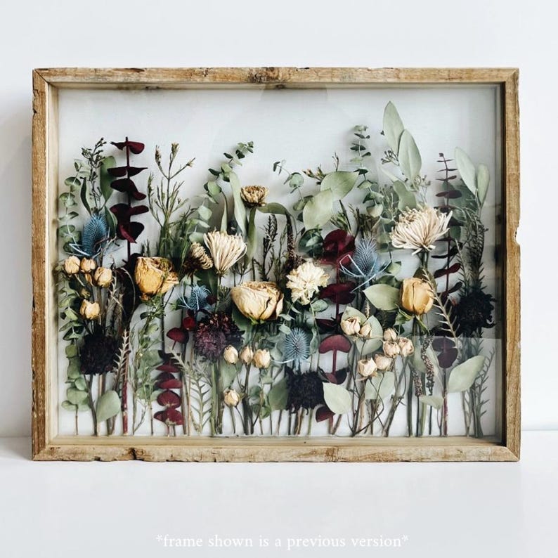 May include: A framed botanical art piece featuring dried flowers and foliage. The arrangement includes yellow roses, blue thistles, and burgundy leaves, set against a clear background. The frame is made of light-colored wood, creating a rustic aesthetic.