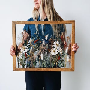 May include: A rectangular wooden framed piece of art featuring a collection of dried flowers and foliage. The arrangement includes white daisy-like flowers, red accents, and various grasses, all encased in clear glass. The frame has a natural wood finish.