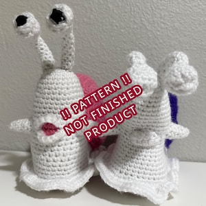 Puede incluir: Dos figuras de caracoles de ganchillo blancos con ojos negros y detalles en rosa y morado. Los caracoles tienen antenas largas y curvas y pequeñas alas blancas. El texto "!! PATTERN !! NOT FINISHED PRODUCT" está superpuesto en la imagen.